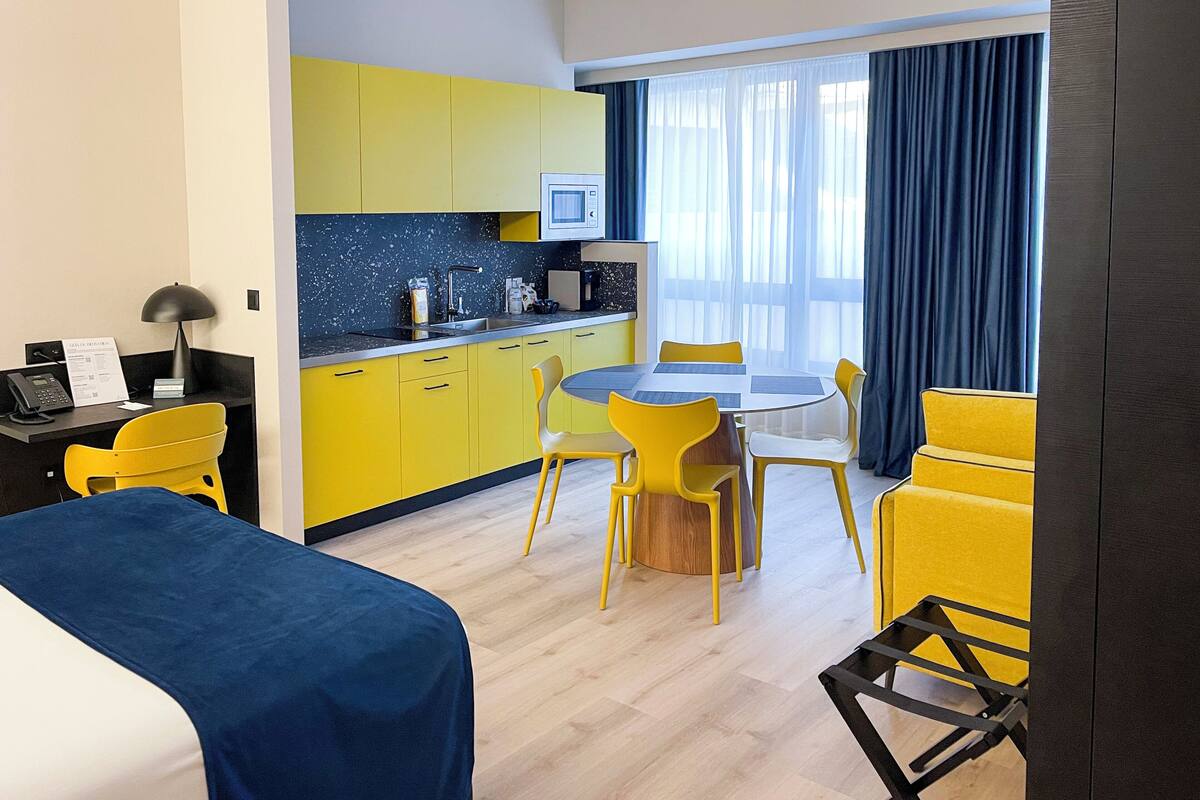 A modern hotel room with a bed, a desk, a kitchenette, and a dining area with yellow chairs.