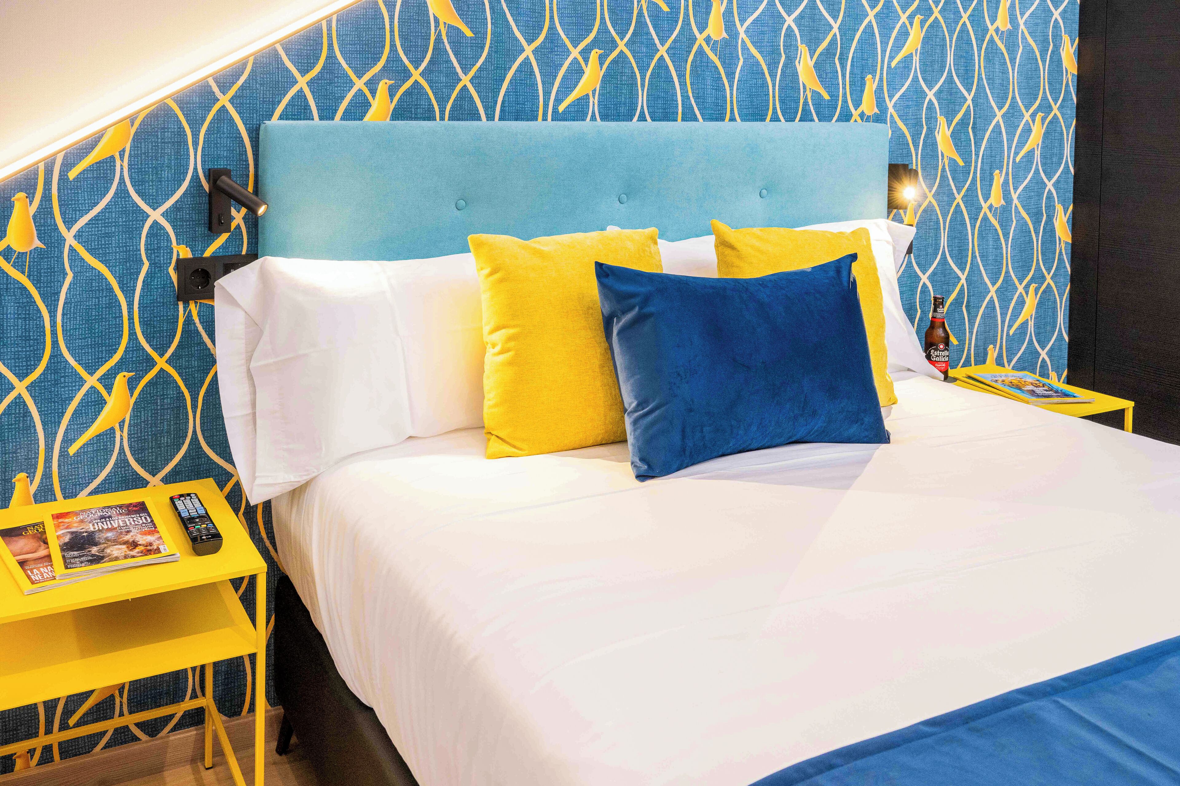 A hotel room with a bed, a nightstand, a remote control, and a magazine.