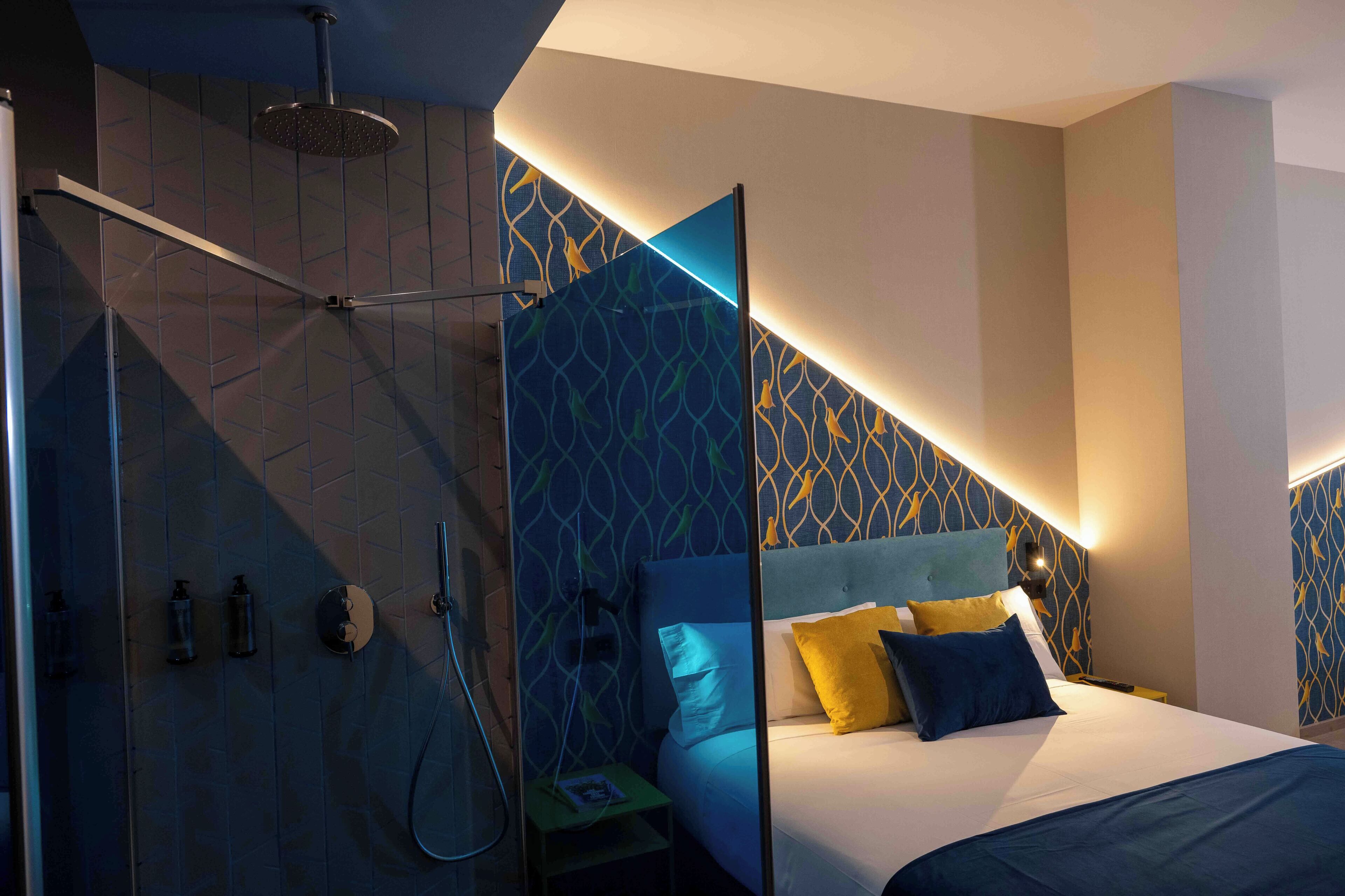 Una habitación de hotel moderna con cabina de ducha de vidrio, una cama con ropa de cama azul y amarilla, y decoración de pared con motivos.