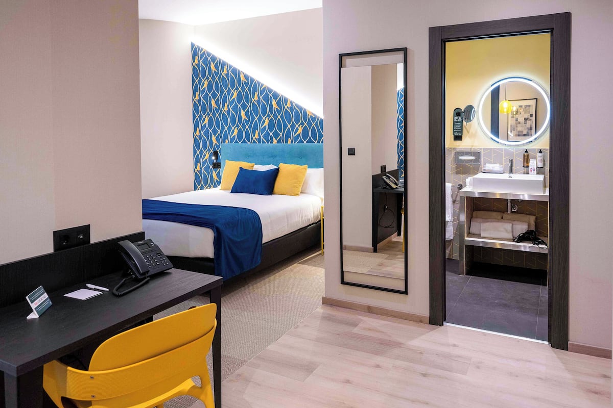 A hotel room with a bed, a desk with a telephone, a chair, and a bathroom with a mirror and sink.