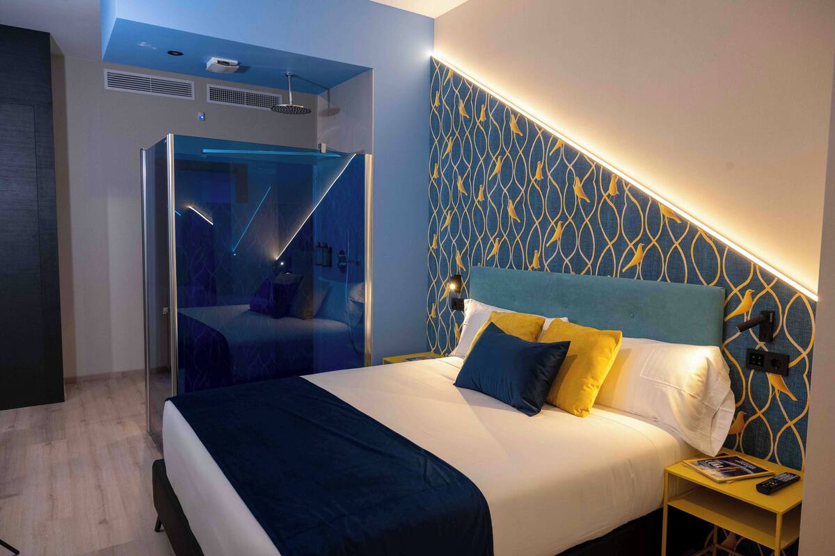 A modern hotel room with a large bed, a glass-enclosed shower area, and a patterned wall.