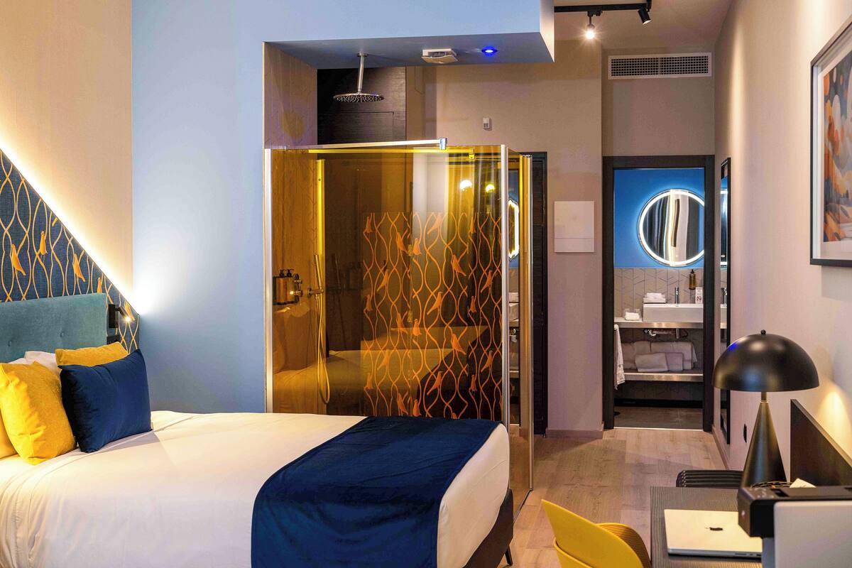 A modern hotel room with a bed, a desk, a chair, a shower area, and a bathroom.
