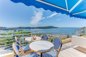 A terrace with a table and chairs, overlooking a coastal landscape with a clear view of the sea and a distant island. - PANORAMIC ESTEREL  AP4475 By Riviera Holiday Homes (Villefranche-sur-Mer)