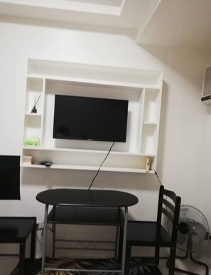 A room with a TV mounted on a white wall, a dining table with chairs, and a fan on the floor.