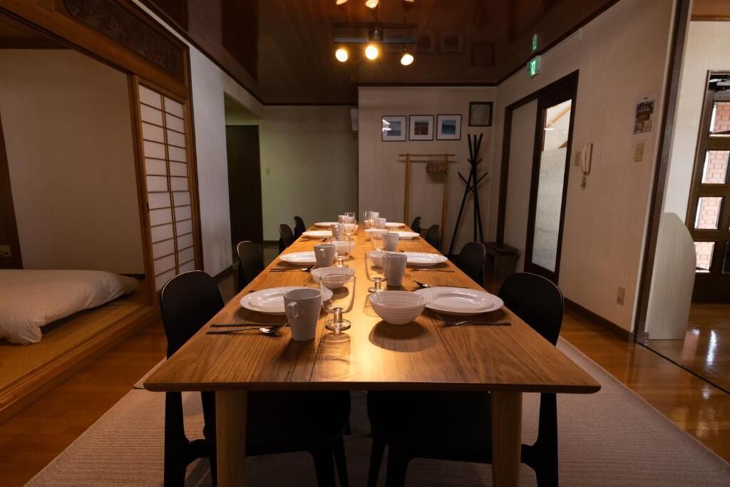 A long wooden dining table set with plates, bowls, and glasses, surrounded by chairs in a room with wooden floors and a traditional Japanese sliding door.