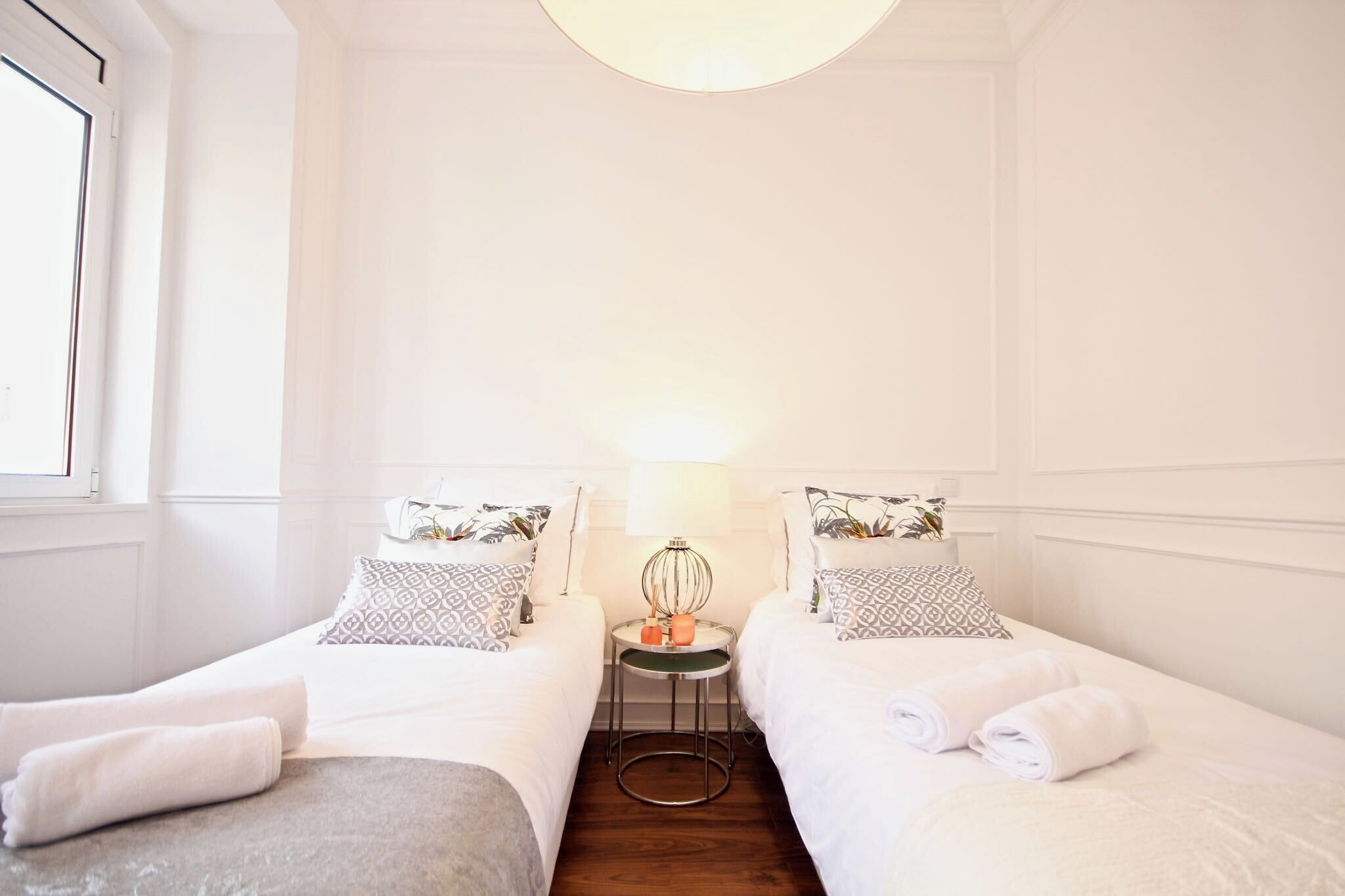Habitación de hotel con dos camas individuales, una mesita redonda con una lámpara y una ventana a la izquierda.