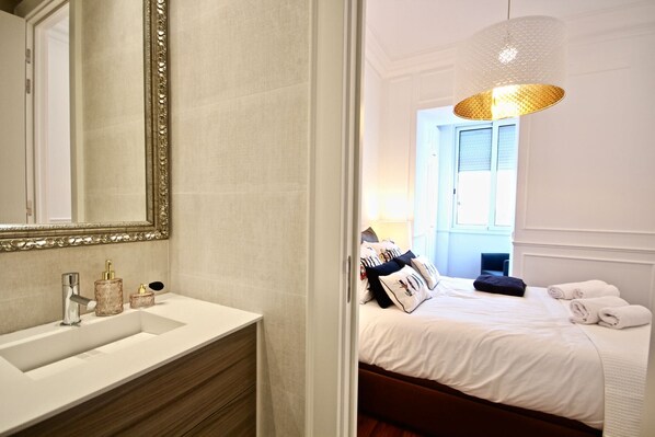 A hotel room with a white vanity, a large mirror, a bed with white linens, a hanging light fixture, and a view of the outside.