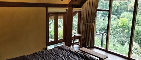 Un dormitorio con una cama grande, un aplique colgante, vigas de madera y vistas a la vegetación a través de amplios ventanales.