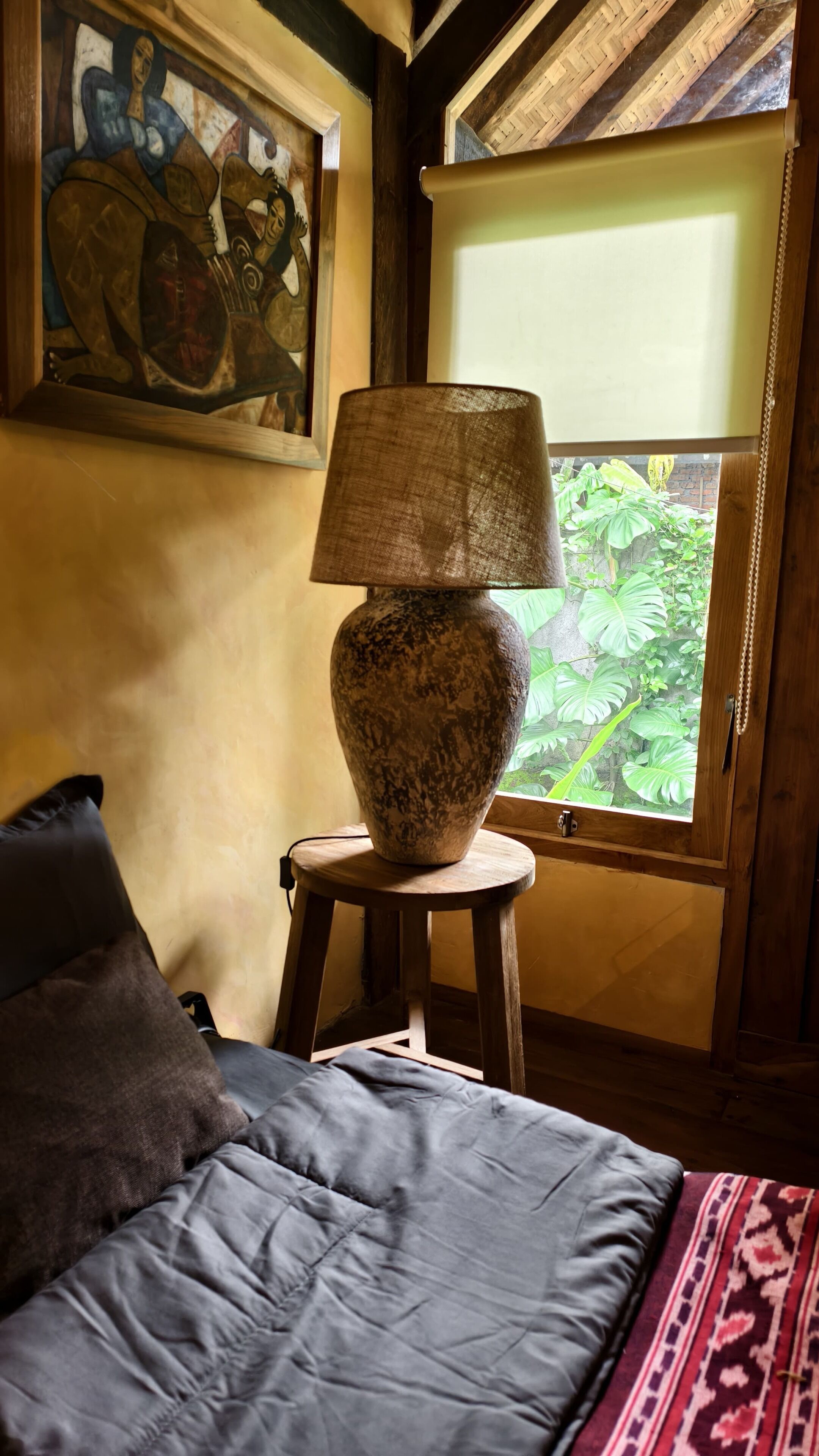 A bedroom with a bed, a lamp on a stool, a painting on the wall, and a window with a view of greenery.