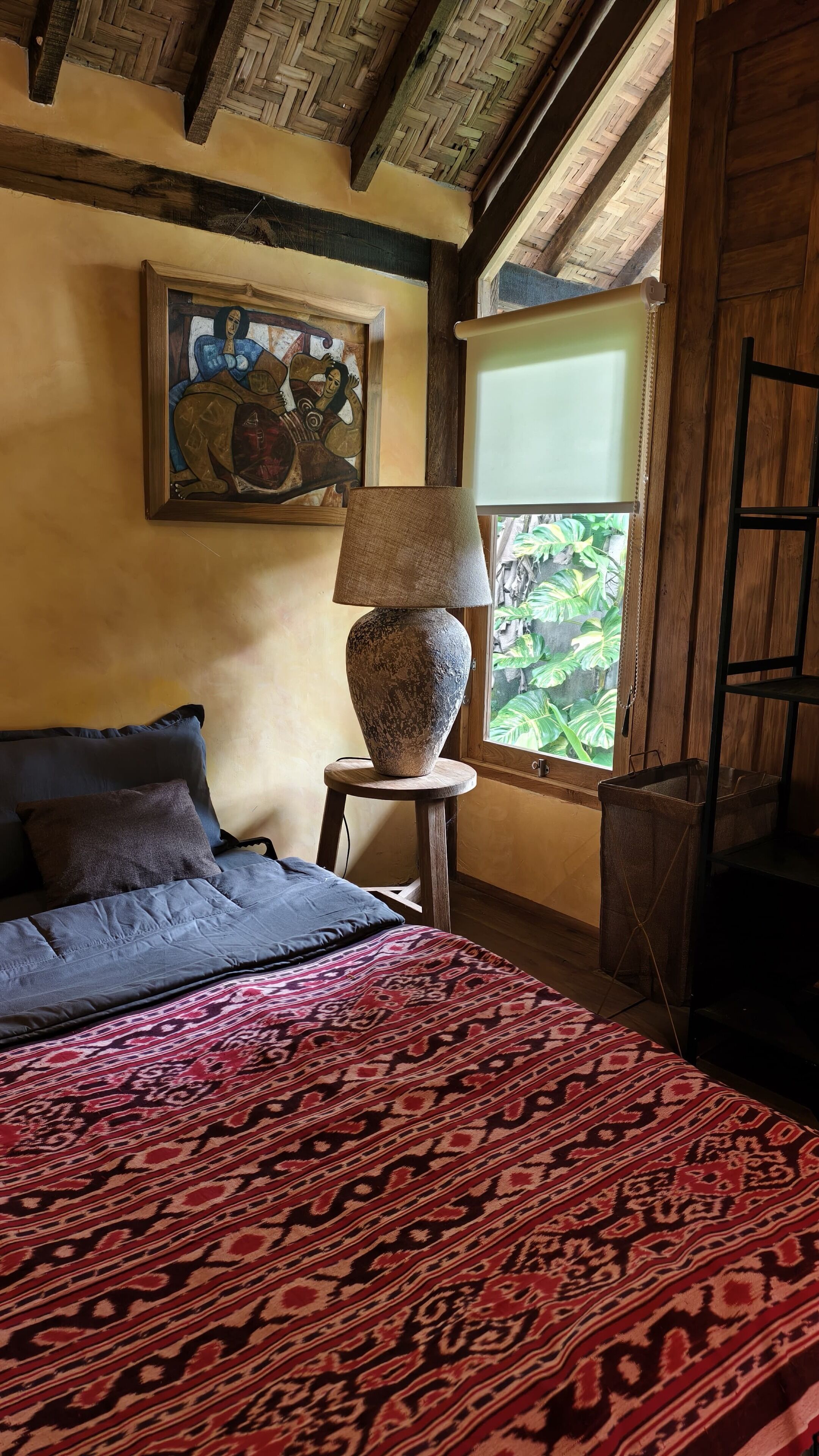 Ein Schlafzimmer mit Bett, einer Lampe auf einem Hocker, einem Bild an der Wand und einem Fenster mit Blick auf Grünflächen.