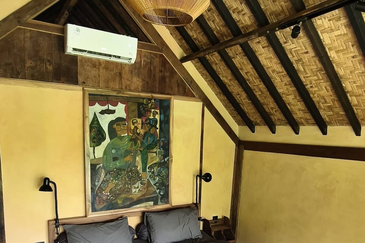 A cozy bedroom with a wooden bed, a bench, a hanging light fixture, and a large painting on the wall.