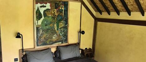 A cozy bedroom with a wooden bed, a bench, a hanging light fixture, and a large painting on the wall.