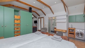 Une chambre avec deux lits, une armoire verte, un banc en bois, un miroir, un lave-linge et un espace douche.