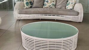 A modern living area with a curved glass coffee table and a white wicker sofa with patterned cushions.