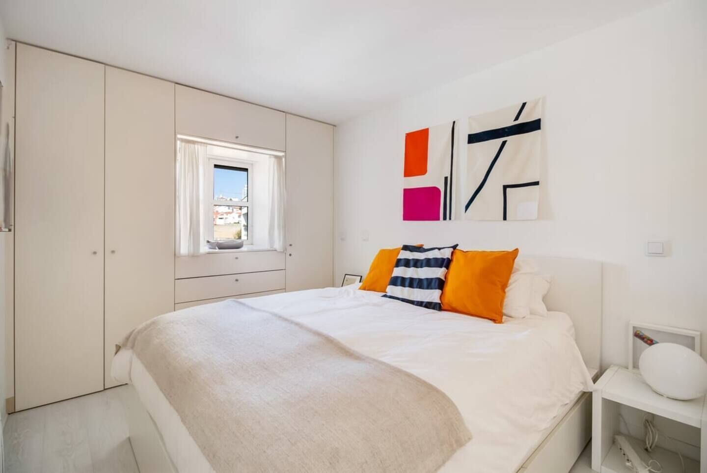 Een slaapkamer met een bed, een raam, een kledingkast en een muur met abstracte kunst.