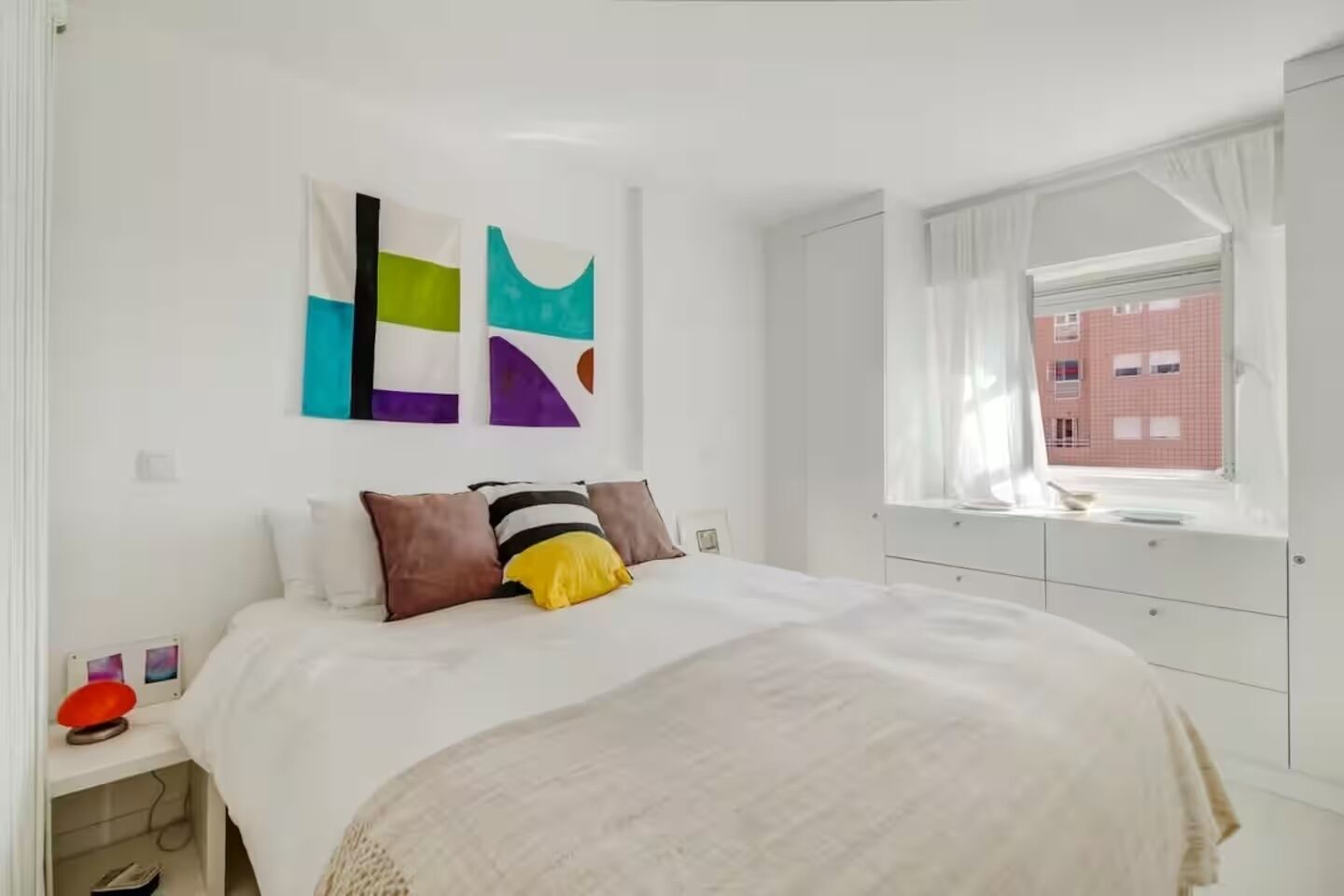 A modern bedroom with a large bed, abstract wall art, a bedside table with a lamp, and a window with a view of a red building.