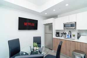 Eine moderne Küche mit weißen Schränken, einem Edelstahlkühlschrank, einer Mikrowelle und einer Kaffeemaschine. Ein Glastisch mit Stühlen und ein an der Wand montierter Fernseher, der 'NETFLIX' zeigt.