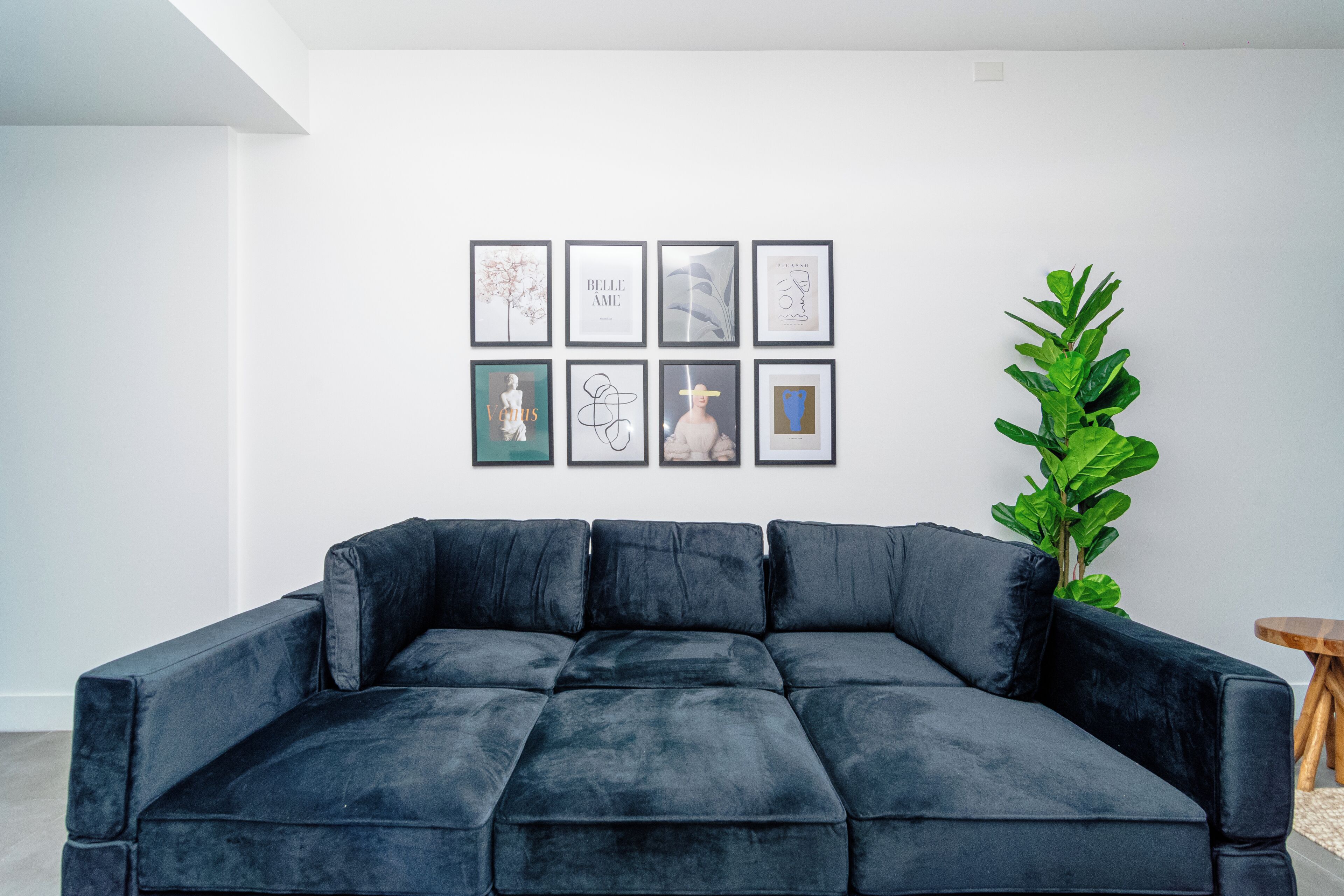 Un salon avec un canapé d’angle bleu foncé, une plante en pot et un mur orné de tableaux encadrés.