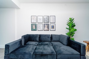 Ein Wohnzimmer mit einem dunkelblauen Ecksofa, einer Topfpflanze und einer Wand mit gerahmten Kunstwerken.