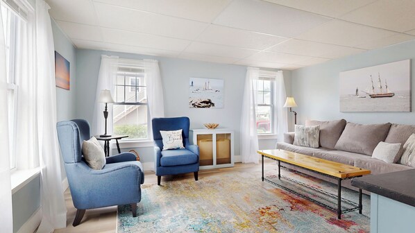 A living room with a blue armchair, a grey sofa, a wooden coffee table, and a colorful rug. There are two windows with white curtains, a lamp, and a small table with a decorative item.