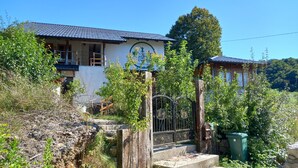 A traditional building with a tiled roof, a wooden staircase leading to a gated entrance, and a lush green garden. - Bosnian house next to the Una river, with a view of the old Ostrovica fortress (Kulen Vakuf)