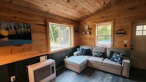 A cozy cabin interior with a sectional sofa, two windows, a painting, and a wall-mounted heater. - Charming cabin in Beautiful Patten Maine with WiFi, ATV/Snowmobile trail access (Patten)
