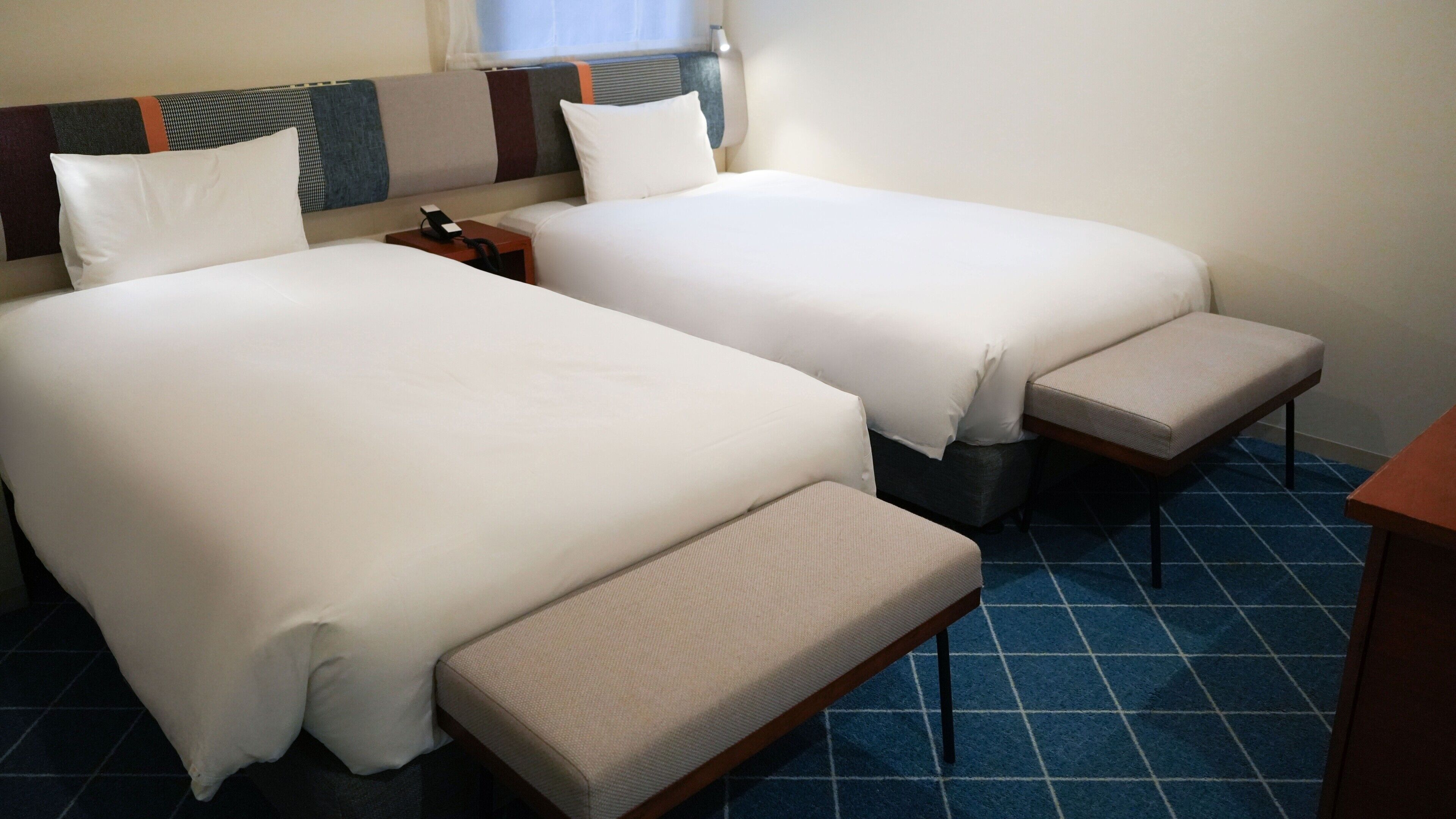 Une chambre d’hôtel avec deux lits, une table de chevet avec un téléphone, un banc et une petite table.