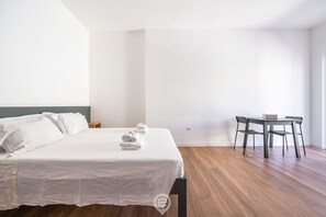 Quarto de hotel com cama, uma pequena mesa e cadeiras, e piso de madeira.
