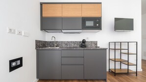A modern kitchen with grey cabinets, a wooden upper shelf, a built-in microwave, a coffee machine, and a TV on the wall.
