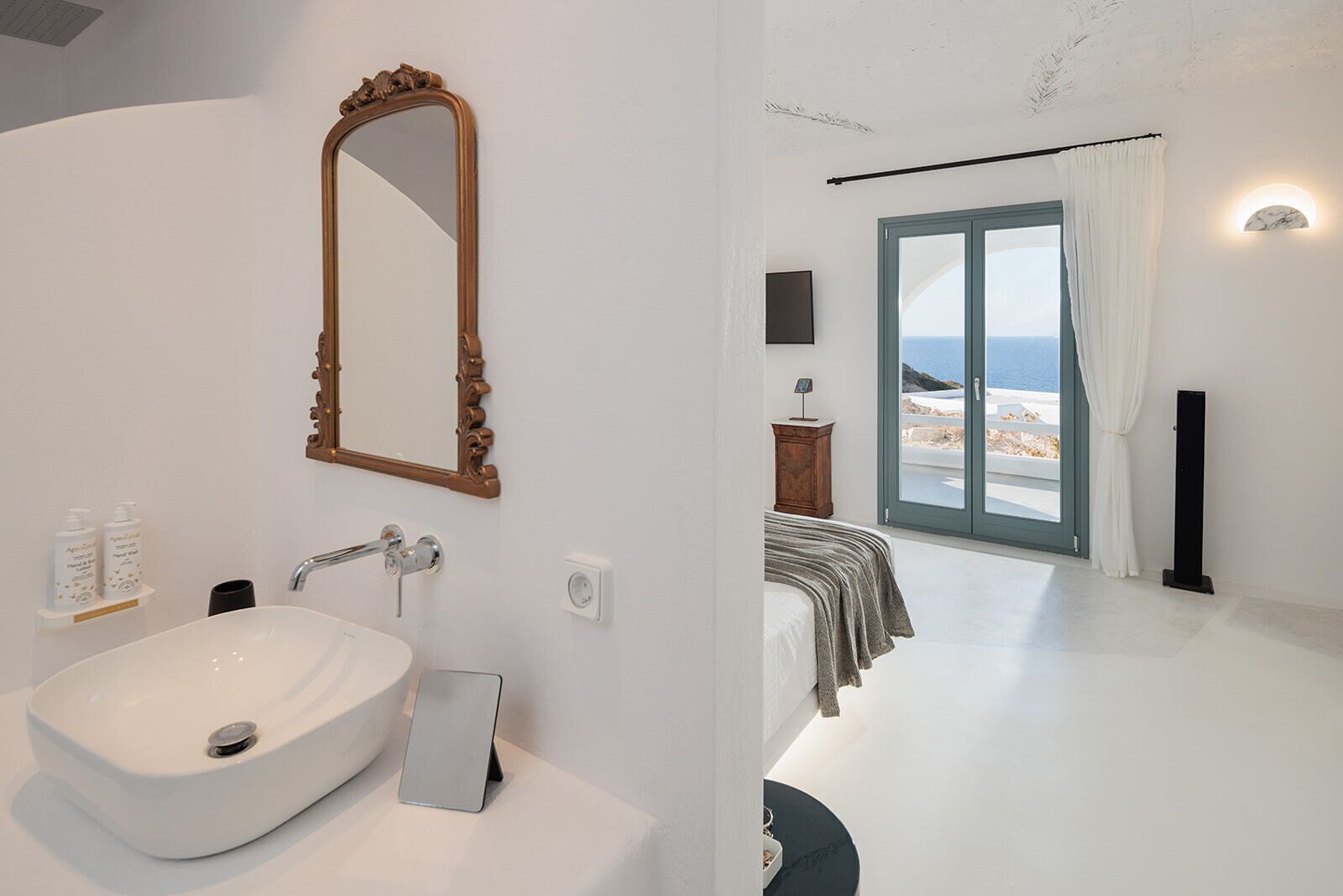 A modern bathroom with a large mirror, white sink, and toiletries. The bedroom has a bed, a nightstand, and a view of the sea through a large window.