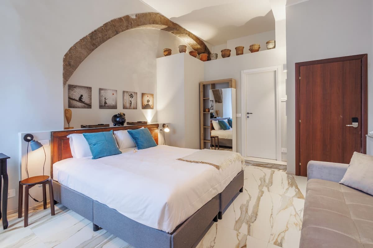 A bedroom with a large bed, a wooden headboard, a small stool, a nightstand, a lamp, and a wardrobe with a mirror.