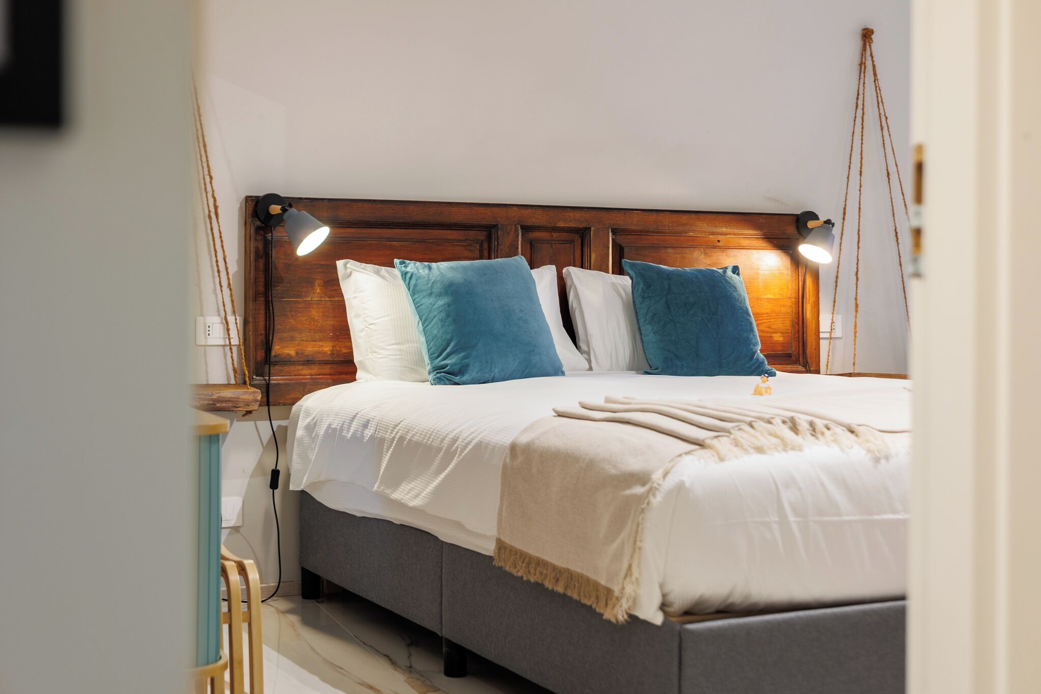 Una cama bien hecha con cabecera de madera, ropa de cama blanca y almohadas azules. Hay dos lámparas de noche a cada lado de la cama.