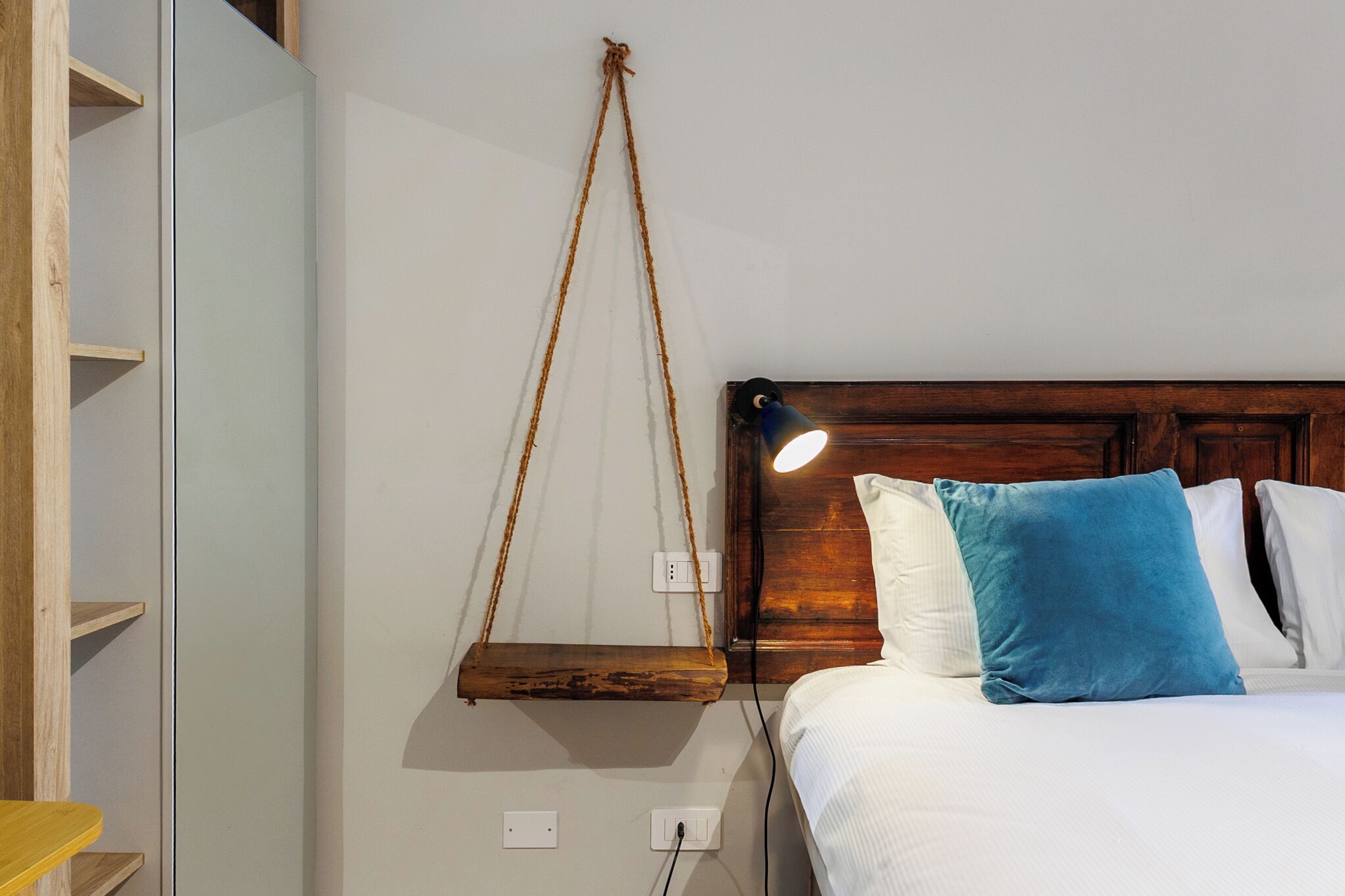 A bed with a wooden headboard, white and blue pillows, and a hanging wooden shelf with a light above it.