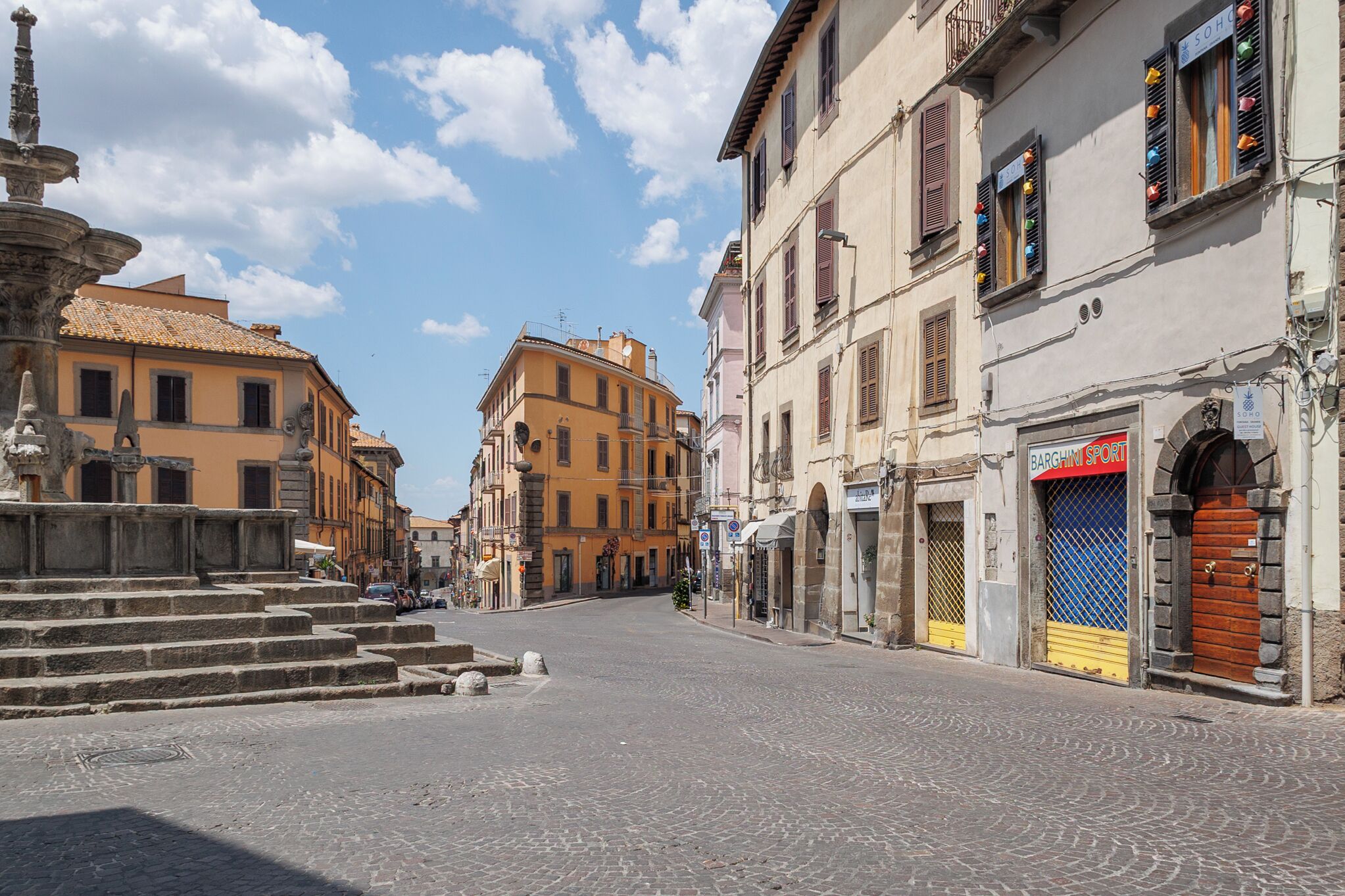 A cobblestone street with historic buildings, a fountain, and a closed shop with a blue and yellow sign.