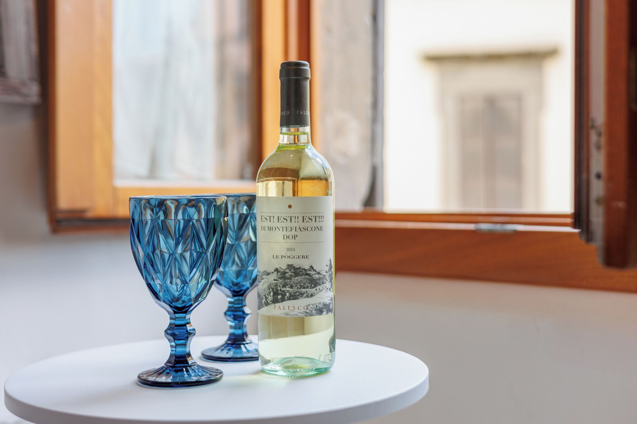 Une bouteille de vin et deux verres en verre bleu sur une table blanche près d’une fenêtre.