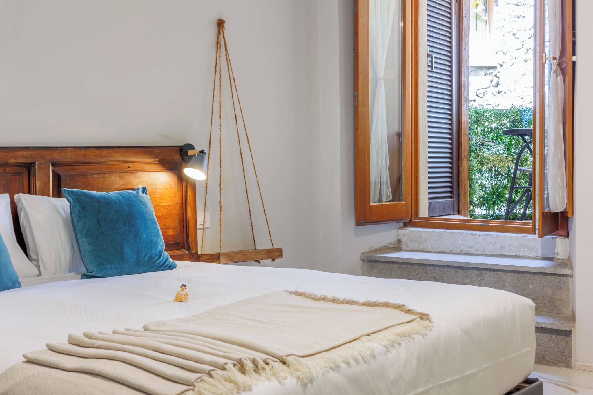 Una cama bien hecha con cabecera de madera, un columpio colgante y una ventana con contraventanas.