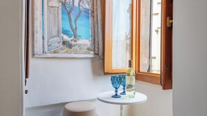 A small room with a painting of a coastal scene, a table with a bottle and glasses, and a window with a view of buildings. - SOHO Fontana Grande By Dasso (Viterbo)