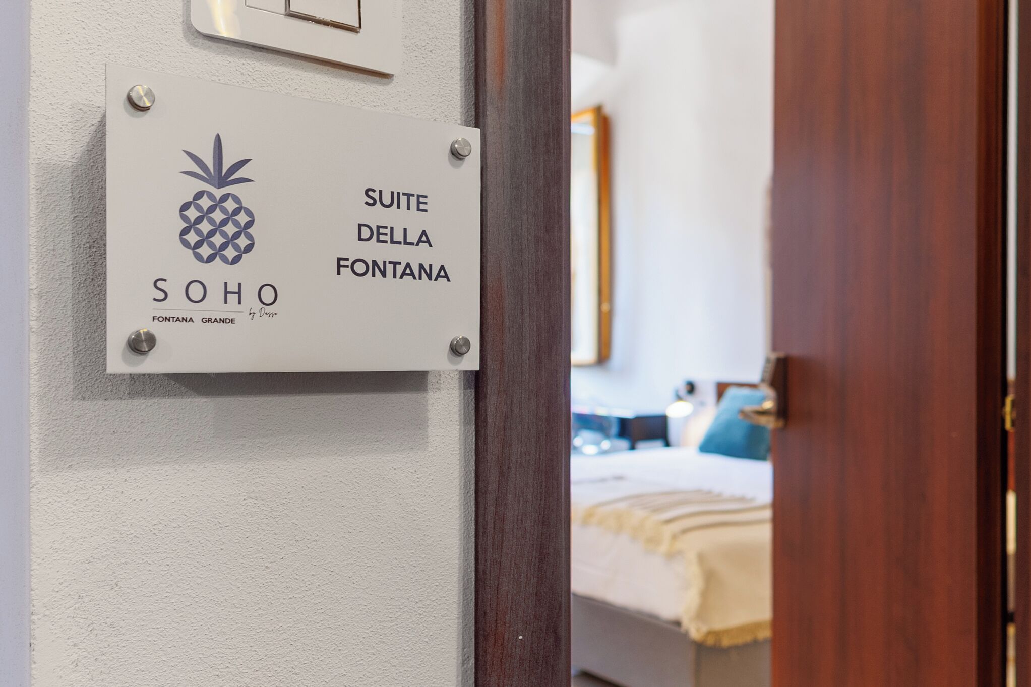 Un segnale che indica la posizione della suite, una porta in legno e uno scorcio di una camera da letto con un letto e un cuscino blu.