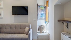 A modern living room with a sofa, a flat-screen TV, a painting of a coastal scene, and a small table with stools. - SOHO Fontana Grande By Dasso (Viterbo)
