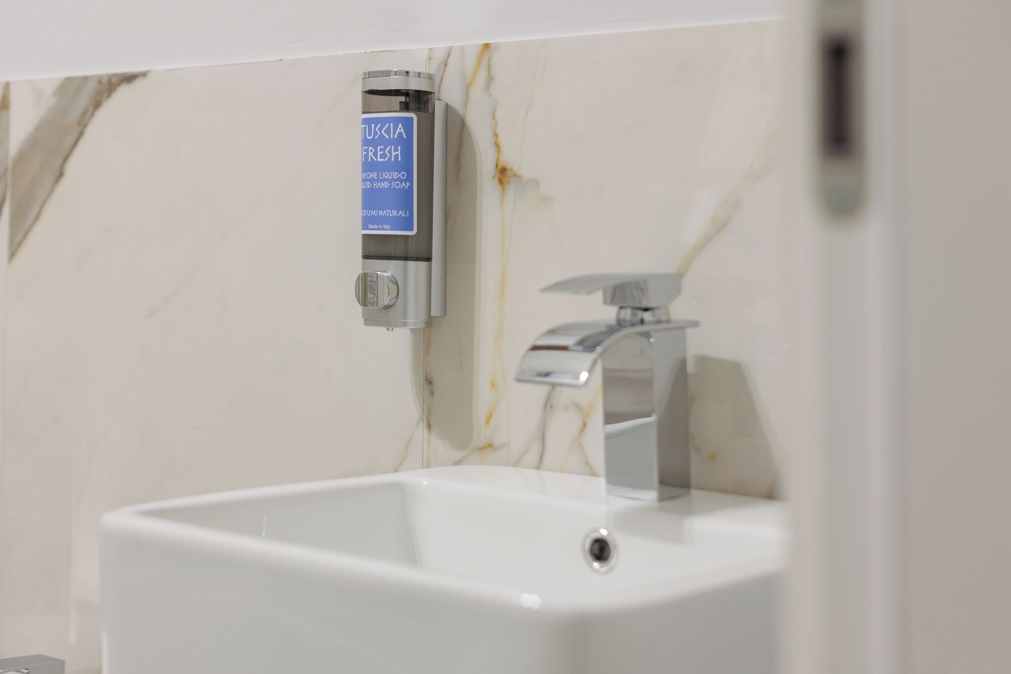 A bathroom with a marble wall, a white sink, and a soap dispenser labeled "TUSKIA FRESH".
