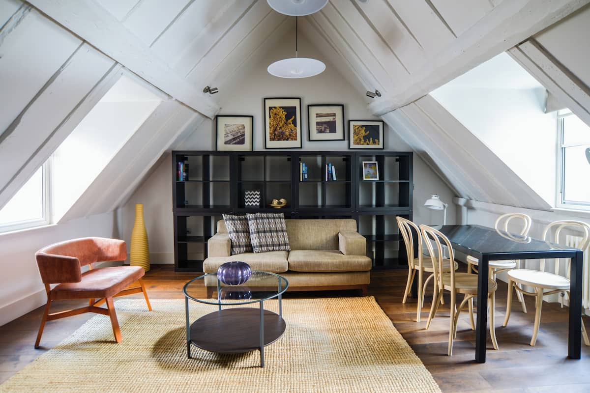A loft-style living room with a high ceiling, a sofa, a coffee table, and a dining area with chairs. There are framed pictures on the wall and a bookshelf.