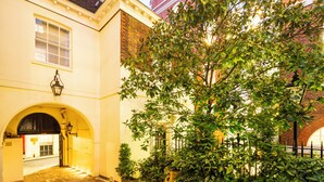 Une cour avec un grand arbre, un bâtiment en briques et une arche donnant sur une rue.