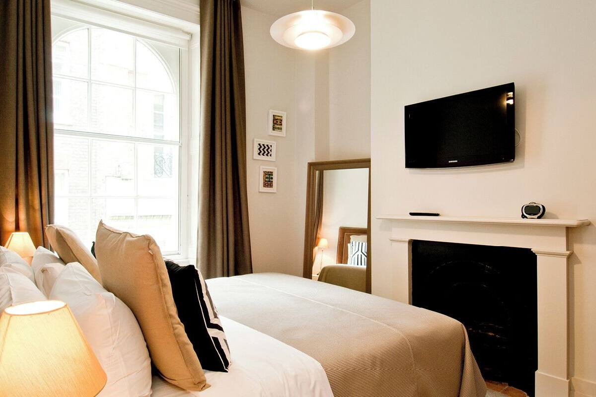 A hotel room with a bed, a TV mounted above a fireplace, and a window with curtains.