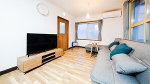 A modern living room with a flat-screen TV, wooden flooring, a grey sofa, and a window with curtains. - Nordic House (Furano)