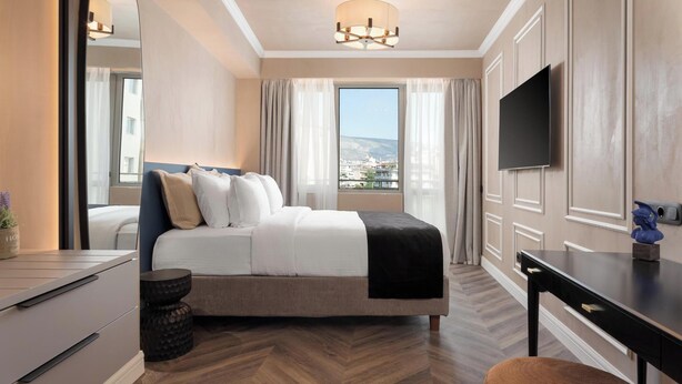 A modern hotel room with a bed, a desk, a television, and a large window offering a city view. - Athena Suite by Acropolis Luxury Athens Suites (Athens)