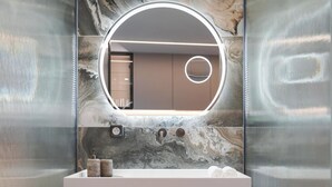 A modern bathroom with a marble backsplash, a round mirror with integrated lighting, a white sink, and two soap dispensers. - Athena Suite by Acropolis Luxury Athens Suites (Athens)