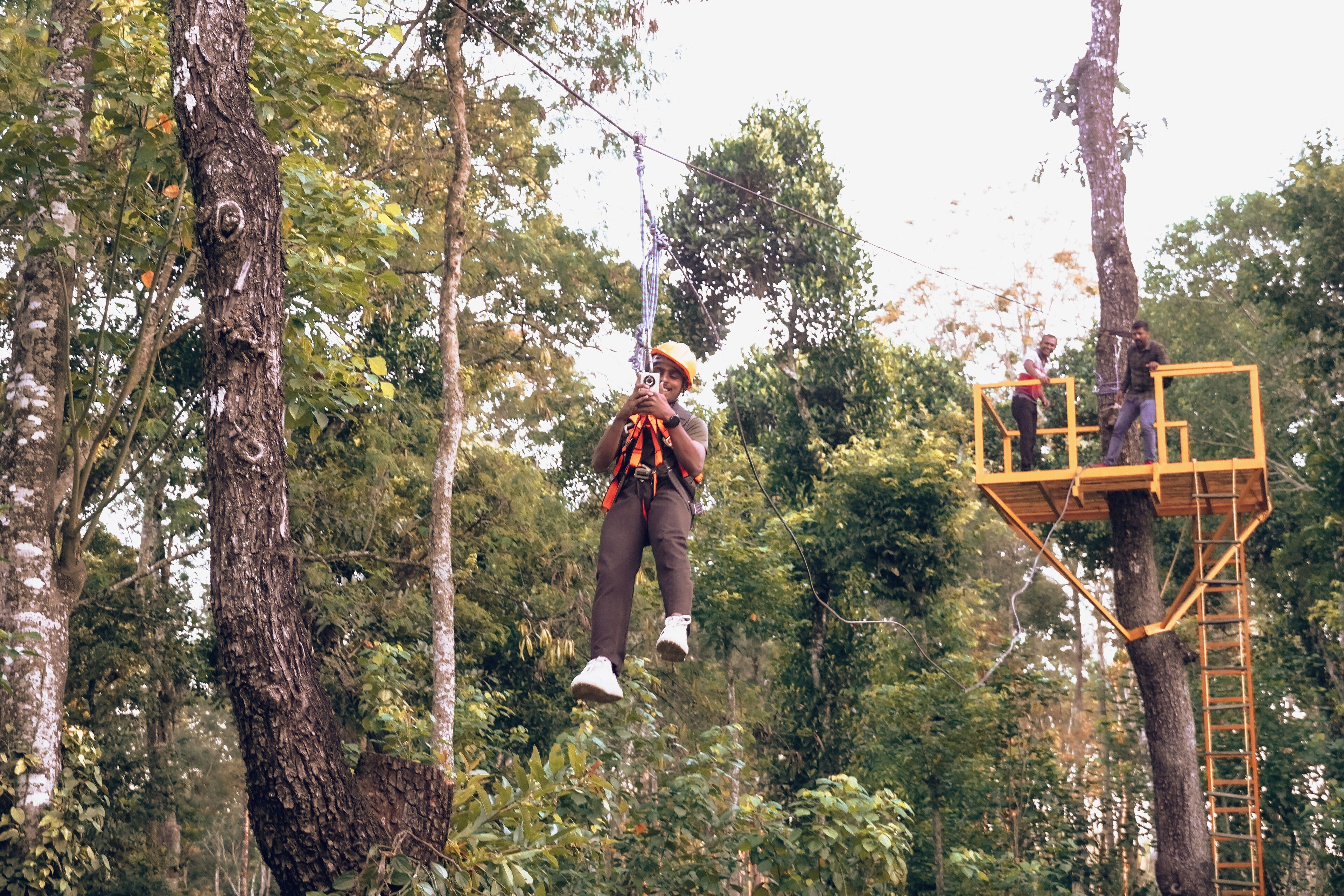 A person suspended in the air, secured with a harness, in a forested area with a wooden platform and ladder.