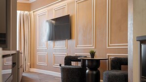 A modern hotel room with a flat-screen TV mounted on the wall, a small round table with a potted plant, and two upholstered chairs. - Aphrodite Suite by Acropolis Luxury Athens Suites (Athens)