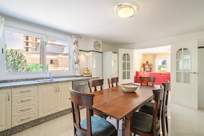 A dining area with a wooden table and chairs, a kitchen with white cabinets, a clock on the wall, and a view of a red sofa and bookshelves in the background. - Beautiful Villa Fortuna for 8 with Private Pool, Terrace & Wi-Fi (El Campello)