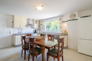 A modern kitchen with white cabinetry, a dining table with chairs, a clock, and a window with curtains. - Beautiful Villa Fortuna for 8 with Private Pool, Terrace & Wi-Fi (El Campello)